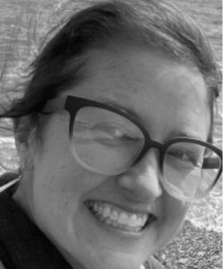 This is a very close-up of a white woman smiling broadly by the sea. She has brown hair and big, two-tone glasses.